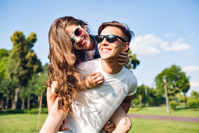 Pretty girl with long curly hair is riding on back of handsome guy in park. They wear sunglasses and have fun. Amore e adolescenza