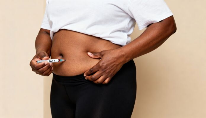 A young Black woman administers an insulin injection into her abdomen. She wears a white shirt and black leggings, focusing on her health management. Farmaci dimagranti: la nuova frontiera che potrebbe aiutare anche a prevenire il cancro