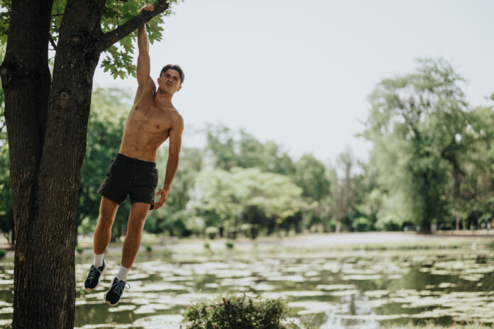 Shirtless man exercising by hanging from a tree branch in a scenic park setting Natural Body Building